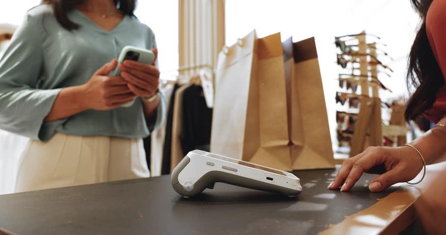 Female, fashion boutique store client paying for clothes using her phone with nfc technology while saleswoman hold card reader machine, close up shot. Transaction, contactless method payment, shopping