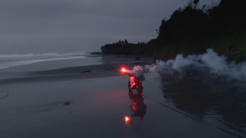 Couple riding a motorcycle on black sand beach at sunset, with a red signal flare, fire. Ocean waves and tropical scenery, romantic vibe. Man, woman driving moto bike on coastline of Bali. Road trip