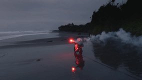 Couple riding a motorcycle on black sand beach at sunset, with a red signal flare, fire. Ocean waves and tropical scenery, romantic vibe. Man, woman driving moto bike on coastline of Bali. Road trip - Powered by Shutterstock - Get 15% off with code: PIKWIZARD15