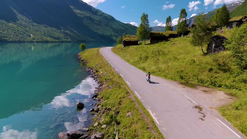 Enjoy the breathtaking beauty of Norways riverside as cyclists glide along a serene road beside crystal-clear waters, framed by lush green hills and majestic mountains of Lovatnet lake Norway