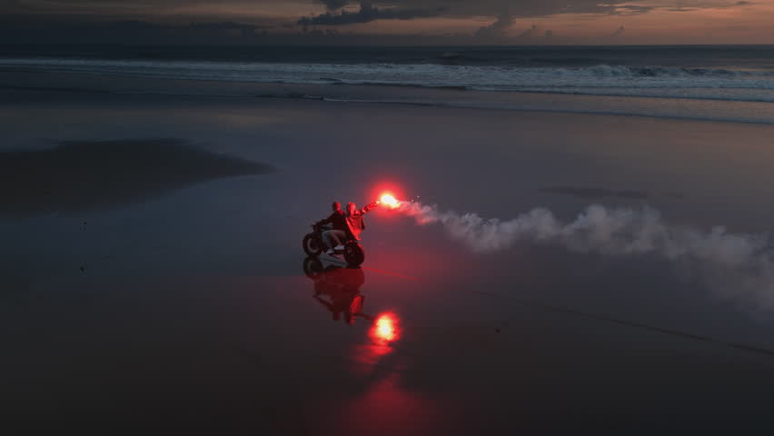 Couple riding a motorcycle on black sand beach at sunset, with a red signal flare, fire. Ocean waves and tropical scenery, romantic vibe. Man, woman driving moto bike on coastline of Bali. Road trip