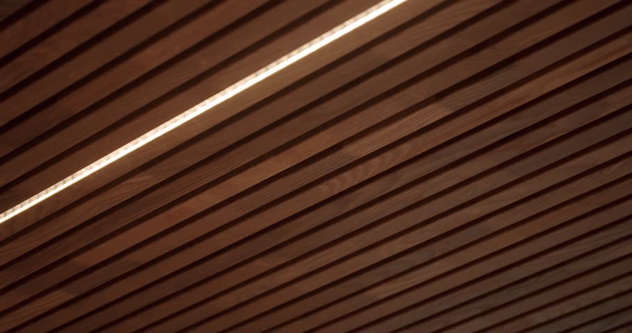 Close-up of wooden slats with a sleek linear light creating a modern, minimalist texture. Ideal for backgrounds and abstract designs. Ceiling with wooden planks and led light lamp at house