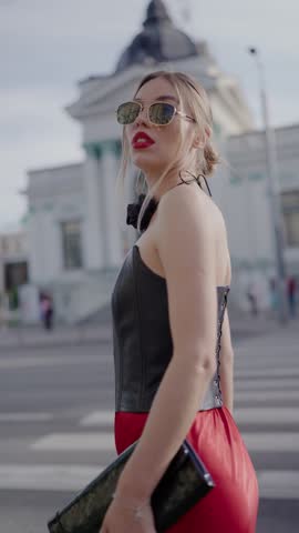 Stylish woman wearing a red dress, sunglasses, and a leather top walking on an urban street. Confident and modern, showcasing chic fashion and city life. Fashionable Woman in Red Dress. Vertical video