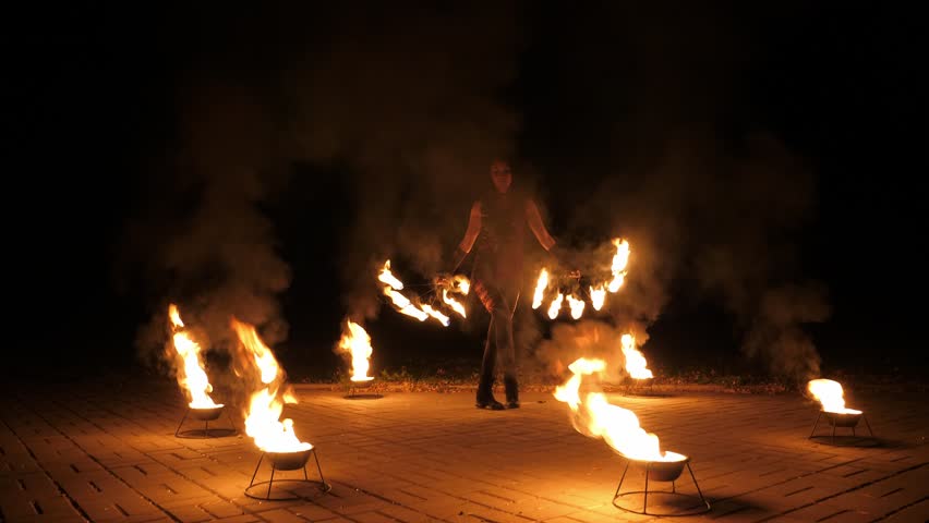 A female stuntman with burning torches bows after a performance in the evening. A woman performs with fire, a professional fire show. There is an inventory for a fire show on the ground.