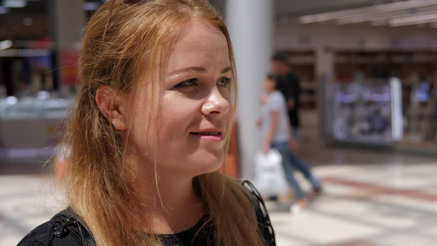 A happy adult woman is drinking coffee in a large bright shopping mall. Portrait of a woman with long blonde hair at the mall, she drinks coffee from a disposable cup.