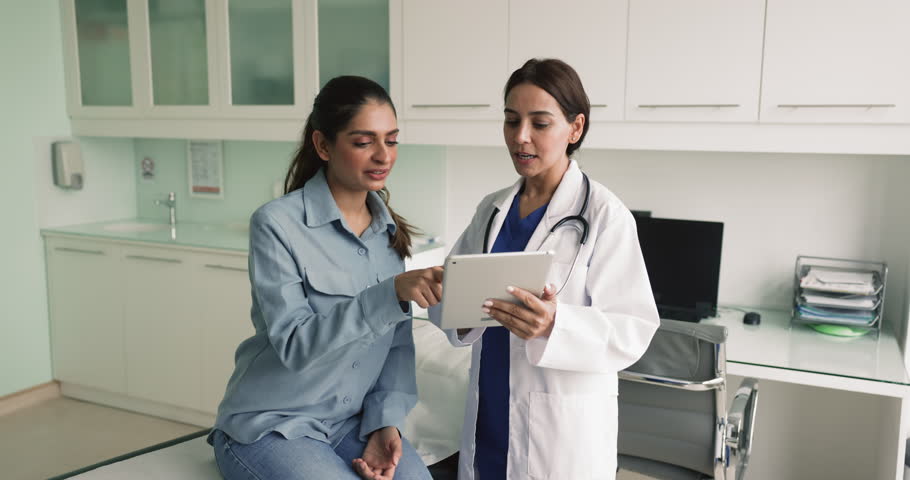 Young Middle Eastern doctor woman talking to female patient in clinic office, showing examination results on tablet explaining image, diagnosis, treatment, recommendation