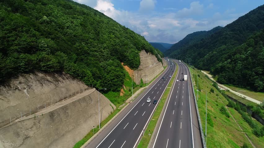 A top-down aerial view of a massive freeway bustling with traffic, weaving through a lush green forest valley, the contrast between the natural landscape.