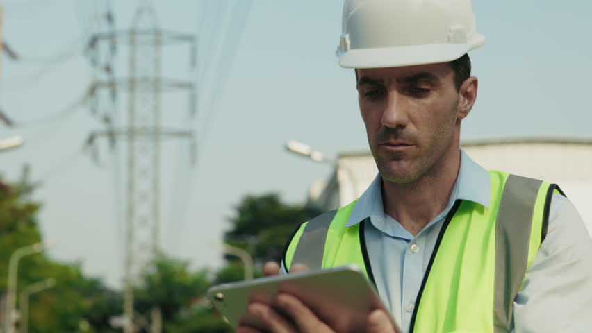 Portrait of man engineer looking away making notes on tablet at construction site. Attentive male builder in helmet and vest checks safety standards examining construction site