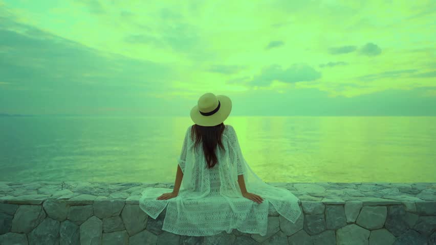 A woman sits on a stone wall overlooking the ocean at sunset. She