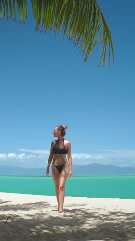 Vertical Screen Video. Girl in bikini walking sand tropical beach, palm trees, clear blue sky. White sand beach, ocean in background. Female relax outdoor lifestyle travel on summer holiday vacation.