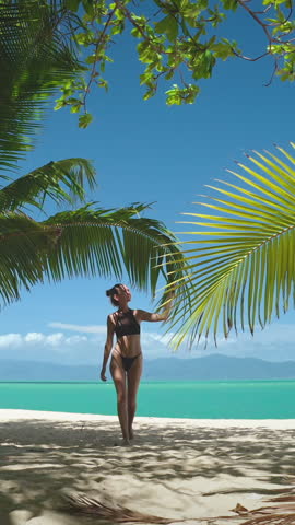 Vertical Screen Video. Girl in bikini walking sand tropical beach, palm trees, clear blue sky. White sand beach, ocean in background. Female relax outdoor lifestyle travel on summer holiday vacation.