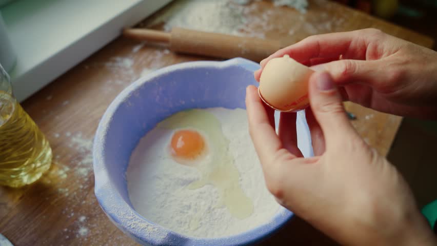 Woman breaking eggshells into bowl filled with flour and egg. Female cracking eggshells over mixture of flour and egg yolk. Lady handling eggshells above baking preparation setup