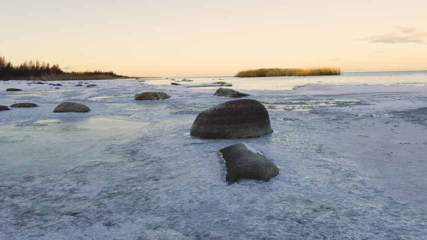 A serene winter scene of a frozen shoreline along Lake Ladoga with scattered rocks partially covered by ice, reflecting the soft hues of sunset in the distance