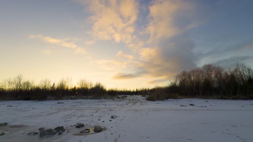 A tranquil winter scene of a snow-covered clearing surrounded by bare trees, with the warm glow of sunset illuminating the sky and soft clouds drifting overhead. Footprints and rocks mark the