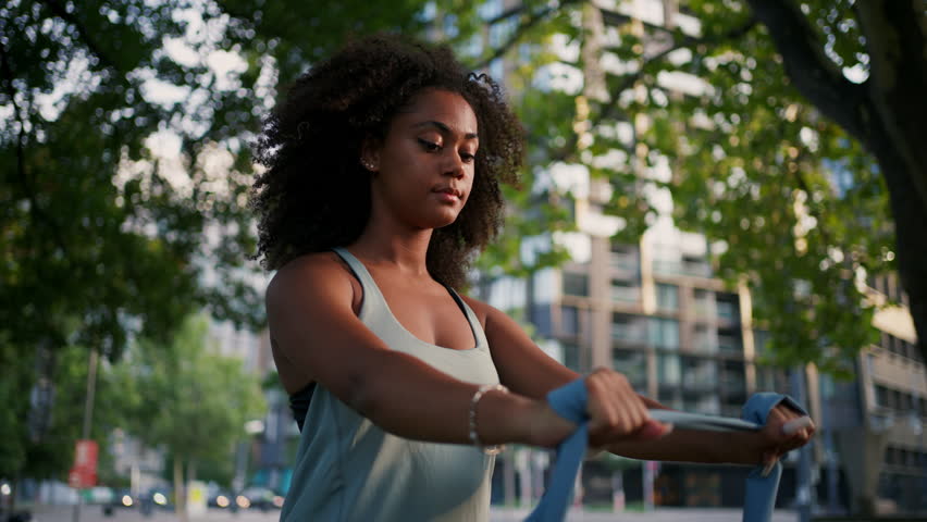 Young sporty woman exercising in city park with resistance band, stretching her arms. Doing pilates outdoors at morning.