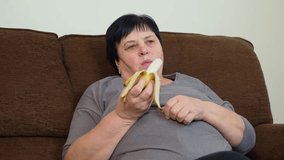 Adult woman sitting on couch eating banana. Senior female relaxing while holding peeled fruit. Lady enjoying snack while resting on sofa - Powered by Shutterstock - Get 15% off with code: PIKWIZARD15