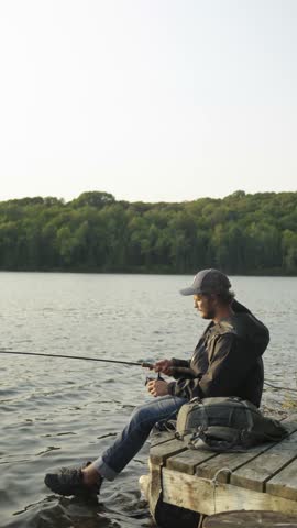 A man wearing a cap and jacket sits on a wooden dock by a calm lake, fishing with a rod, surrounded by a serene forest landscape in the background