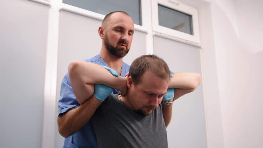 Neck manipulation, Manual therapy, Chiropractic healing. Male patient, seated with his hands behind his head, receives neck adjustment from chiropractor in blue scrubs and gloves.