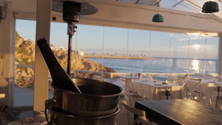 Empty seaside restaurant bathed in sunset light during the golden hour. A bottle of champagne or cava in an ice bucket near the table. Mediterranean interior, terrace, local cuisine, Sitges.
