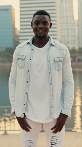 Positive African American guy wearing casual clothes stands on city embankment. Smiling man holds hand on hips standing against downtown commercial buildings. Urban fashion. Vertical shot