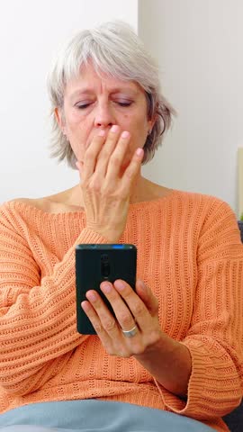 Vertical HD video. Worried senior woman reading bad news on smart phone message sitting on sofa at home.