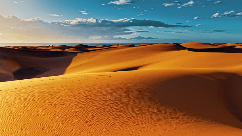 Golden sand dunes stretch across the landscape of Gran Canaria, Spain, illuminated by warm sunlight under a clear blue sky and scattered clouds.