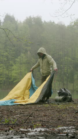 Vertical video. Hiker assembles campsite tent in the autumnal forest near the lake. Traveler installing tent in woods. Outdoor weekend activity. Wide shot