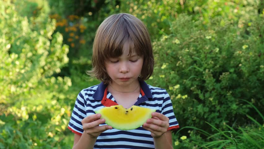 Close up portrait of little boy with long hair holds slice of yellow watermelon in hands and eats it. Happy child enjoys eating fruit in nature. Concept of children's joy, healthy and nutritious food