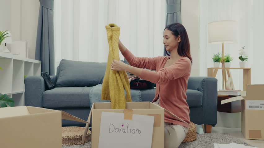 Asian woman carefully packs a yellow sweater into a donation box at home, promoting generosity, sustainability, and community support through charitable giving.