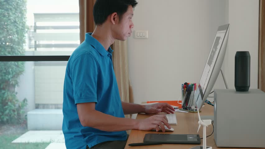 A man is sitting at a desk with a computer monitor and keyboard in front of him. He is wearing a blue shirt and he is focused on his work