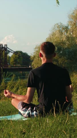 Back view of young man sitting on yoga mat in lotus position and meditating in city park. Male practicing yoga outdoors in sunny day. Yoga morning routine. Vertical video