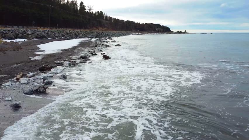 A truck camper drives on a gaspe peninsula coastal road along a snowy beach where small scattered pieces of ice are flattered by the waves of St Lawrence river, Quebec, Canada, 2024.