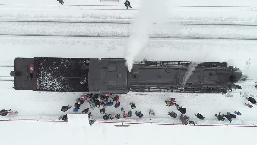 Train is seen in the snow with a large crowd of people gathered around it. The train is black and has a lot of steam coming out of it. The people are standing in the snow. Nizhniy Novgorod