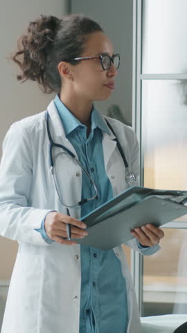 Vertical shot of female doctor in white coat and glasses standing in medical office, commenting on chest x-ray and talking to patient during health consultation