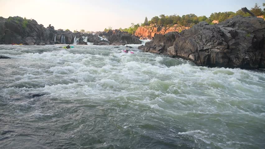 Two men kayaking on a river with rocky banks down a mountainous rough water at sunset.