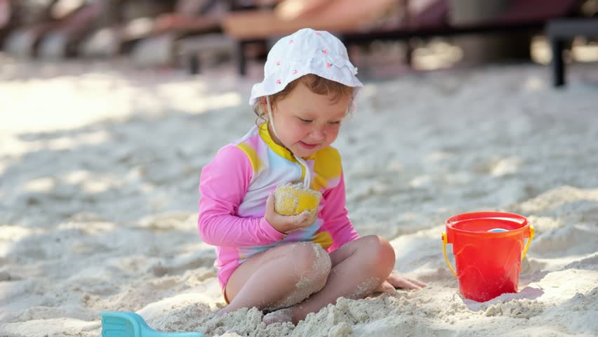 Little girl in pink long-sleeved t-shirt plays in sand on beach. Toddler child sculpts sand figures using plastic molds and bucket on vacation at tropical resort