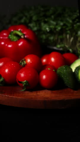 fresh vegetables covered with water rain drops in dark room,rotating on plate.microgreens,tomatoes cherry,cucumber and red paper in dark scene.professional video shooting for advertising restaurants