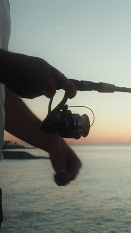 Vertical video. Close-up of a fisherman spins the reel on a spinning the fishing rod against of sunset