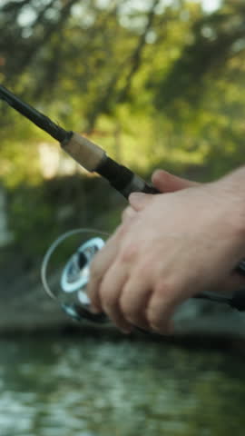 Vertical video. Close-up of man with a fishing rod stands on the shore and enjoying fishing spins the reel on a spinning the fishing rod