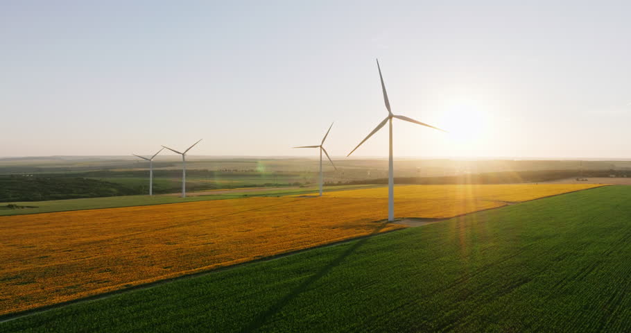 Wind turbines with blades in field aerial view bright orange sunset blue sky wind park slow motion drone turn. Silhouettes windmills, large orange sun disc summer lens flare. Alternative energy