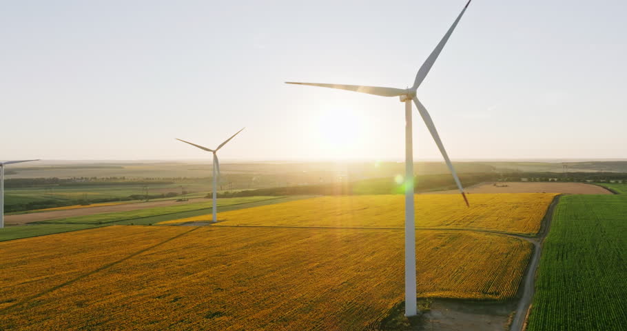 Large wind turbines with blades in field aerial view bright orange sunset blue sky wind park slow motion drone turn. Silhouettes windmills, large orange sun disc summer lens flare. Alternative energy
