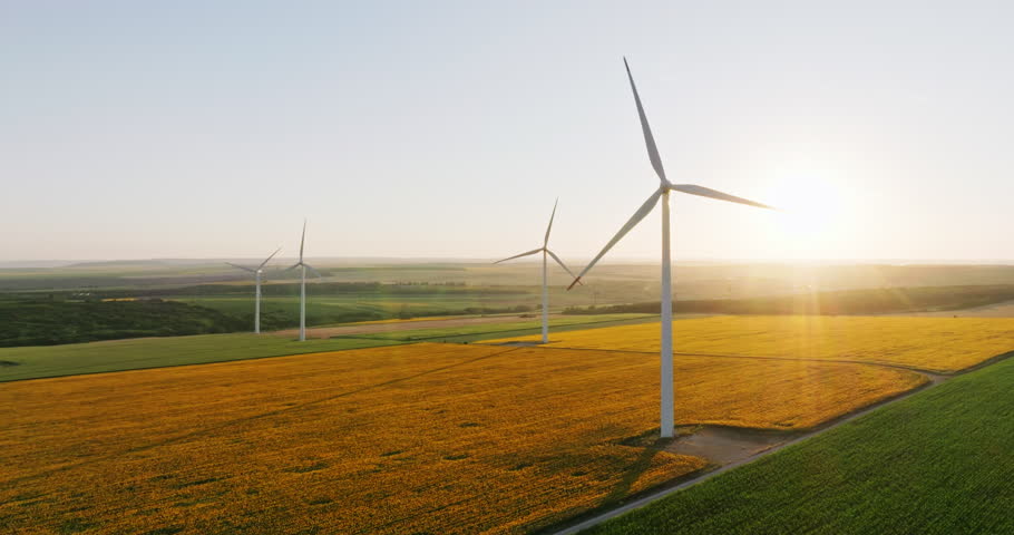 Large wind turbines with blades in field aerial view bright orange sunset blue sky wind park slow motion drone turn. Silhouettes windmills, large orange sun disc summer lens flare. Alternative energy