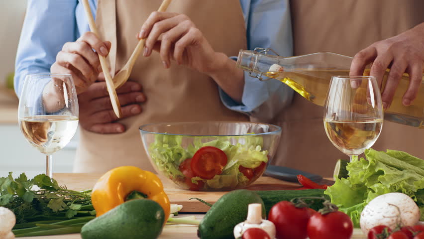 In a cozy kitchen, a couple enjoys preparing a fresh salad. One person pours olive oil into a bowl while the other stirs leafy greens, surrounded by colorful vegetables and wine glasses.