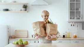 A woman with short hair smiles as she carries two brown paper bags filled with groceries. Fresh produce, including green vegetables and fruits, is visible in the bags and on the counter. - Powered by Shutterstock - Get 15% off with code: PIKWIZARD15