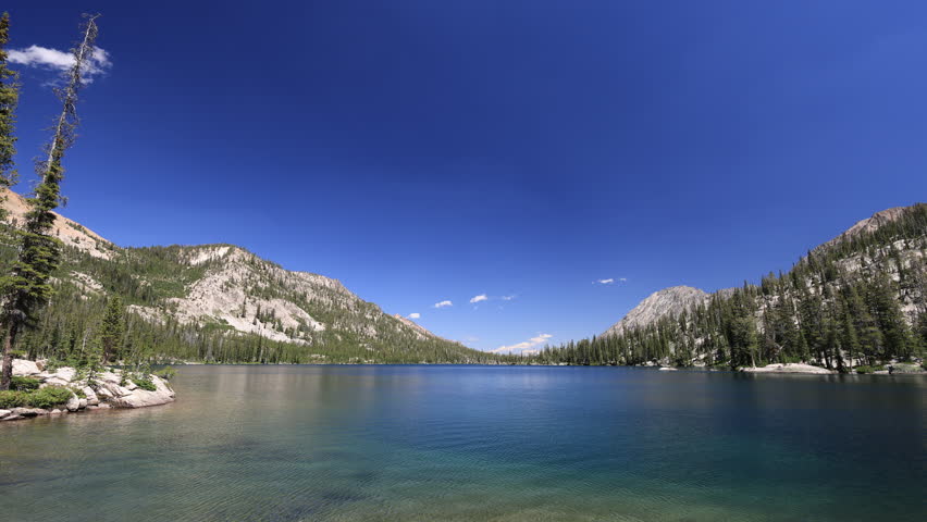 Toxaway Lake, a large alpine lake in the Sawtooth Mountains near Stanley, Idaho, is showcased on a beautiful summer day with a tranquil water surface under a blue sky and a few distant clouds.
