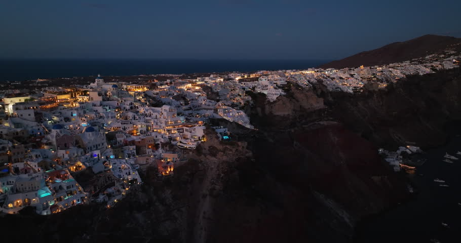 Aerial high angle view of beautiful town of Oia on the steep cliff of Santorini Island, Greece. Illuminated white buildings at dusk with boats on the ocean
