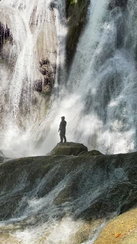 A man stands near a majestic waterfall, watching clean water cascade into a serene river in the mountains.