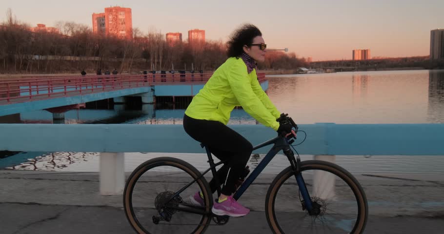 A middle aged woman in a neon jacket rides a bicycle along a riverside path at sunset, embracing an active and healthy lifestyle