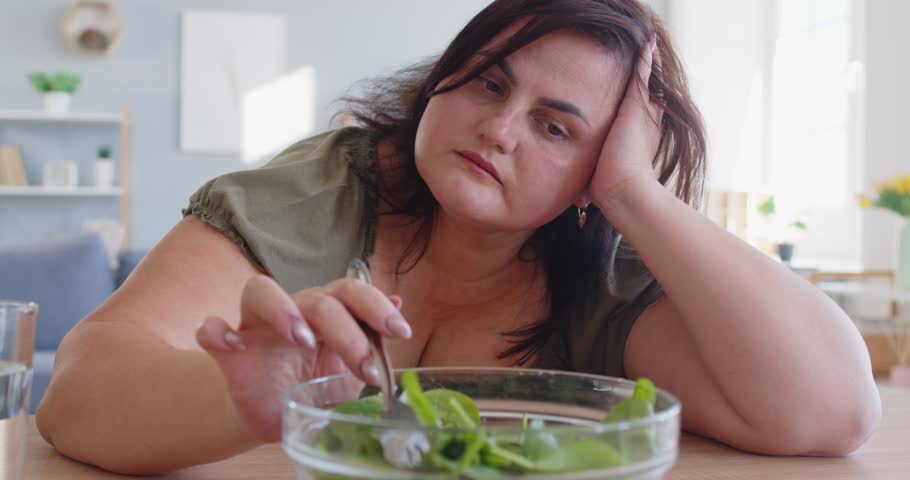Portrait of unhappy sad fat woman sitting at table in kitchen in front of a bowl of green salad. Overweight girl eating healthy food at home. Nutrition, dieting and weight loss concept. 4k video.