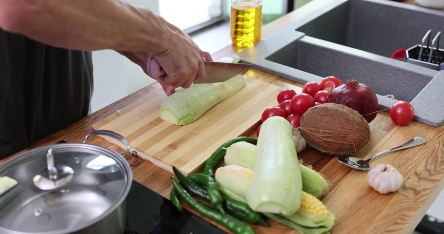 Man chef cuts zucchini with knife on wooden board in kitchen at home. Male cook prepares vegetarian dinner standing at modern countertop slow motion
